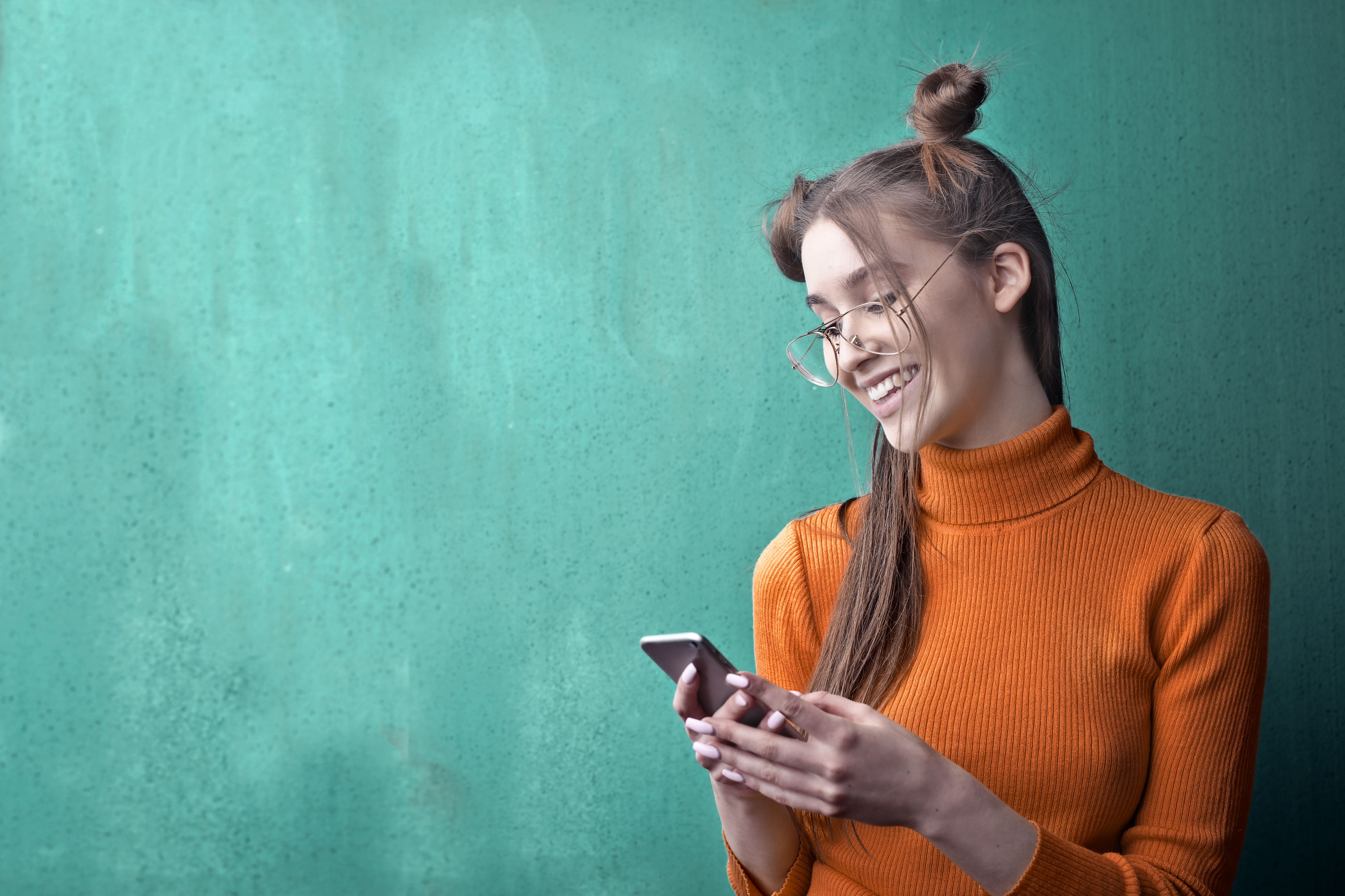Una mujer con gafas y jersey naranja consulta su móvil delante de una pared verde; imagen decorativa.